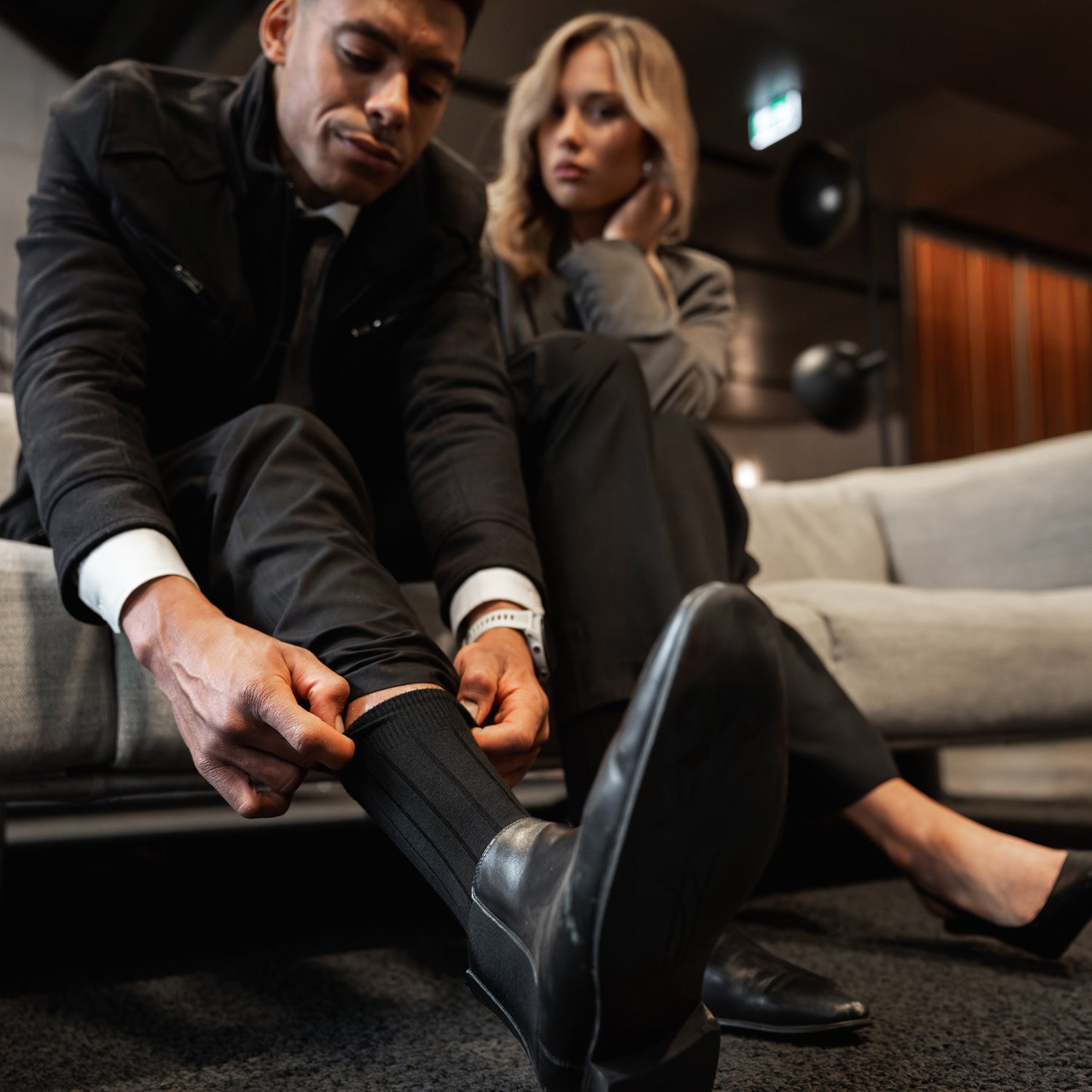 Mann im Anzug zieht schwarze Businesssocken von BLACKSOCKS hoch, sitzt neben Frau im Business-Look auf Sofa.